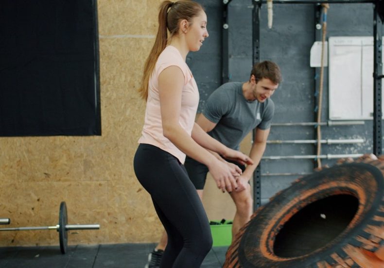 beautiful young woman training in gym with rubber tyre lifting weight working out with trainer. People, sports workout and sports concept.
